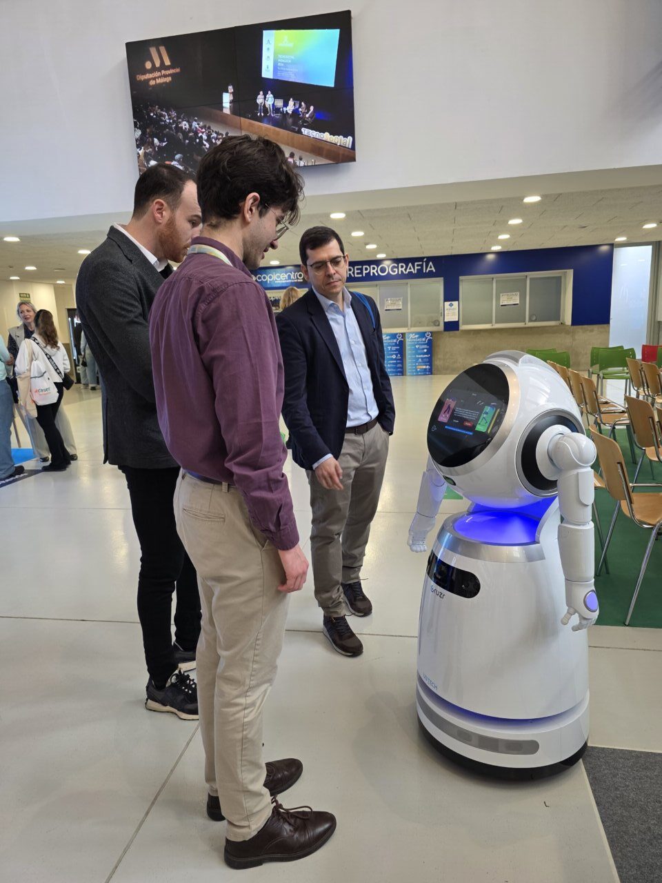 Tres personas observan un robot en un congreso sobre servicios sociales.