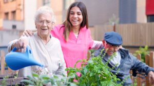 Mujer mayor riendo mientras riega plantas con ayuda de cuidadora y anciano