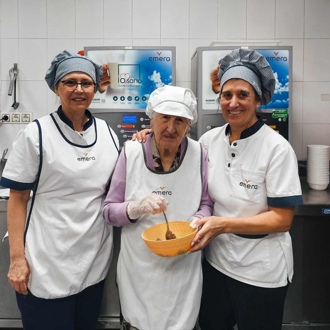 Tres personas en una cocina, una anciana y dos trabajadoras de Emera cocinando juntas.