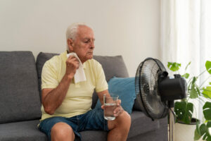 Hombre mayor sentado en un sof&aacute; con un vaso de agua y un ventilador