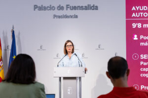 Una mujer habla desde un atril ante asistentes y un panel sobre ayudas públicas.