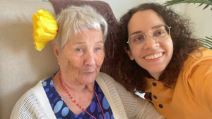 Una mujer mayor sonriente con una flor amarilla en su cabello junto a una joven.