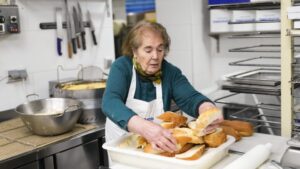 Mujer mayor coloca trozos de pan en una bandeja dentro de una cocina.